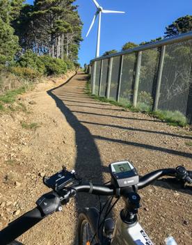 Point of view from an electric mountain bike, looking towards the handle bars. The bike is facing towards an uphill dirt track with a large white wind turbine in the distance.