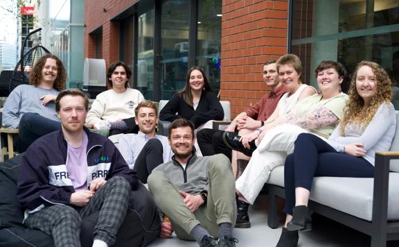 A group of ten people sit and smile together on a exterior patio in an urban setting.