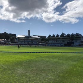 The Basin Reserve looking towards the grandstand. It’s sunny and a cricket game is underway on the field.