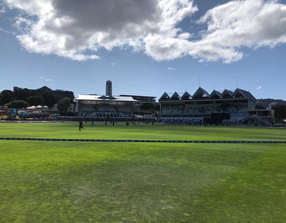 The Basin Reserve looking towards the grandstand. It’s sunny and a cricket game is underway on the field.