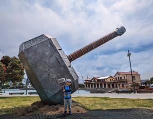 A child wears a Thor costume, holding a prop hammer, is in front of the Thor Hammer, Mjölnir statue located at the Whairepo Lagoon on Wellington Waterfront.