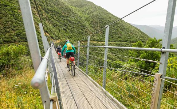 2 people biking across a swing bridge over a valley in the Remutaka Range, on the Remutaka Cycle trail.