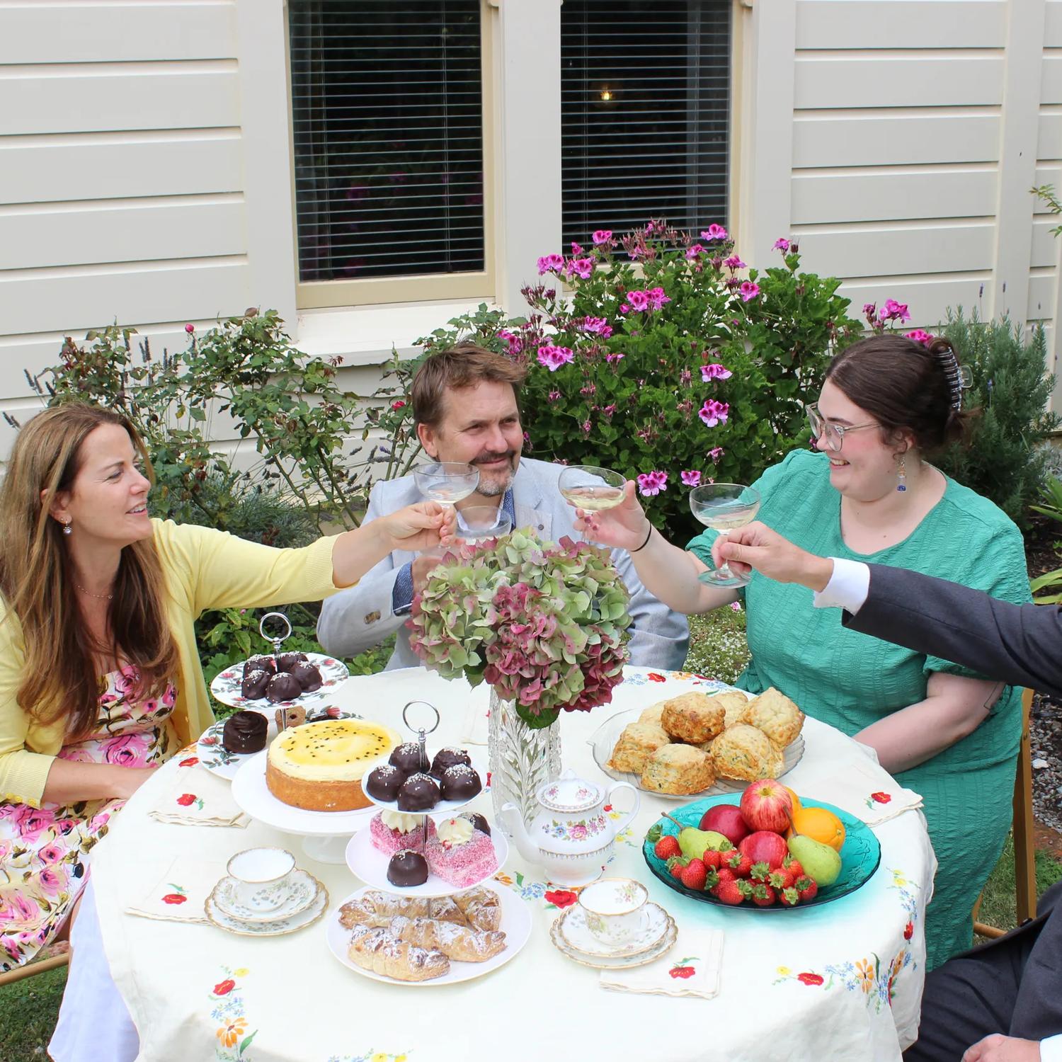 Four people make a toast with coup glasses and sit around a round table with fruits, desserts and a flower vase.