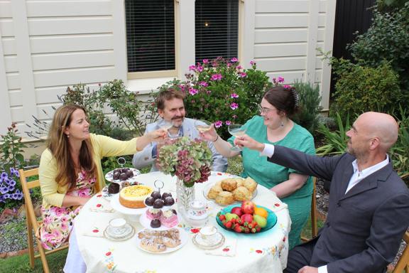 Four people make a toast with coup glasses and sit around a round table with fruits, desserts and a flower vase.