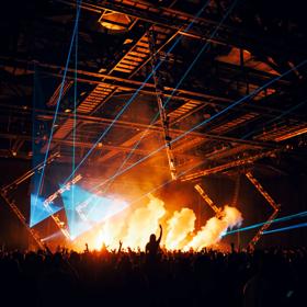 A packed crowd of people inside a large dimly lit venue space with blue laser lights and orange coloured smoke clouds.