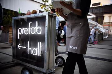 A caterer from Nosh walks past an LED sign that says 'Prefab hall'.