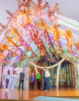 Eight people standing on stage below Rongomaraeroa - the marae at Te Papa which is covered in ornate colourful traditional maori carvings.