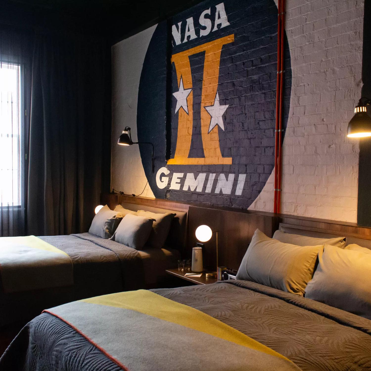 Inside a room in The Intrepid Hotel in Wellington, with two double beds and brick wall is painted with a large circle, inscribed with 'NASA' at the top and 'Gemini' at the bottom. The Gemini symbol, is centrally placed within the circle.