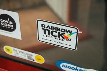 Close up of a white Rainbow Tick Certified sticker with black text and a rainbow checkmark on a glass pane.