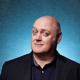 Dara Ó Briain, wearing a dark blazer and shirt, posed against a textured blue background, gazing directly at the camera.