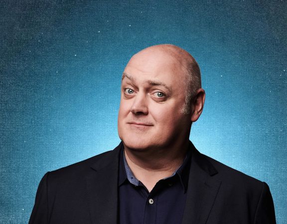Dara Ó Briain, wearing a dark blazer and shirt, posed against a textured blue background, gazing directly at the camera.