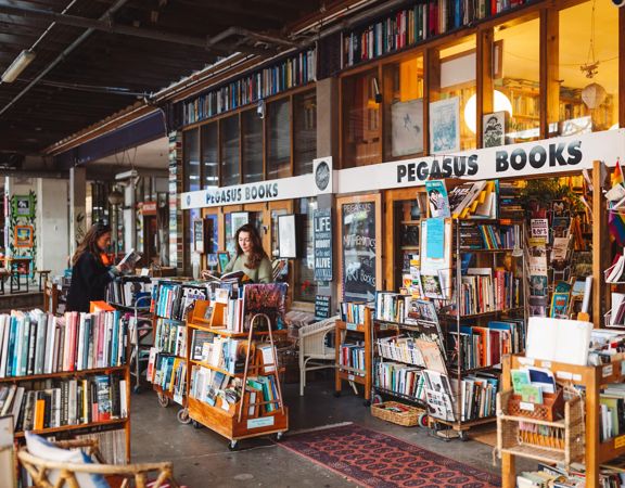 Two people browse through books outside Pegasus Books in Left Bank off Cuba Street. Pegasus Books is an older wooden building with shelves of eclectic books outside.