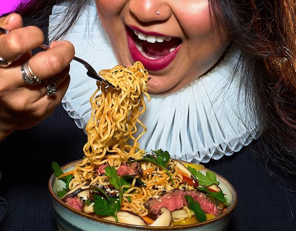 A close-up shows a person holding a bowl of noodles and lifting a forkful to eat, with greens and slices of meat visible in the dish.