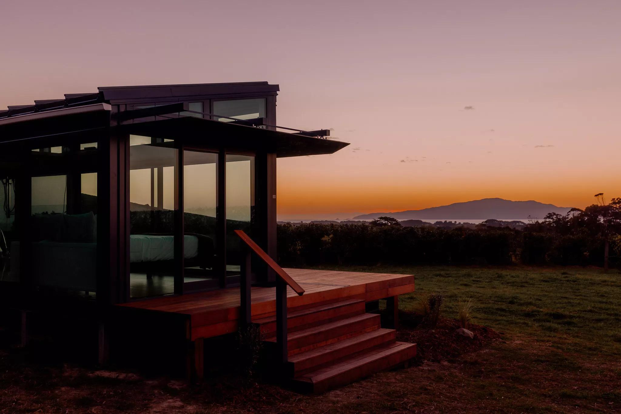 Kokomea PurePod is a luxurious glass eco-cabin that sits on a grapevine nursery high above the Kāpiti Coast. The sky is a gradient of hazy pink to bright orange on the horizon with Kapiti island seen in the background. 