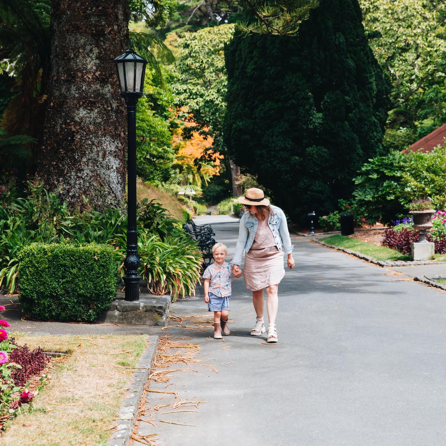Mother and child walking down a concrete path surrounded by leafy trees and flowers.