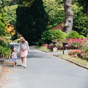 Mother and child walking down a concrete path surrounded by leafy trees and flowers.