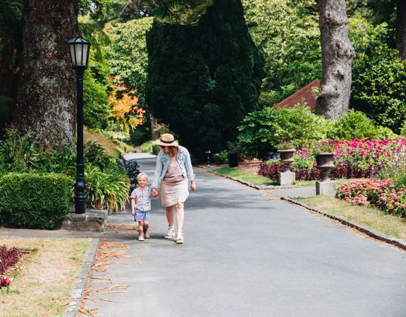 Mother and child walking down a concrete path surrounded by leafy trees and flowers.