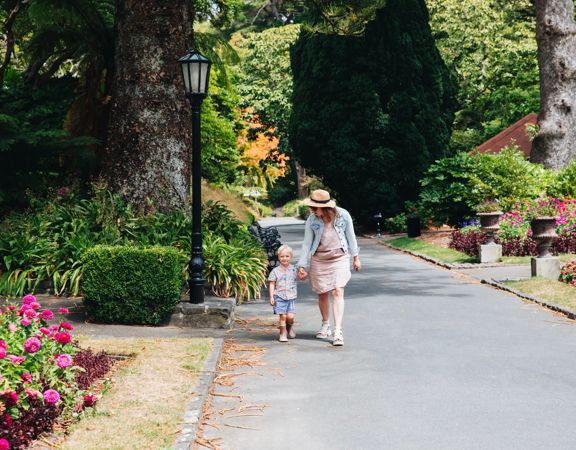 Mother and child walking down a concrete path surrounded by leafy trees and flowers.