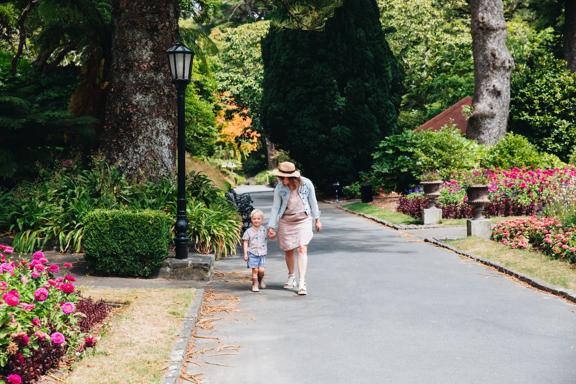 Mother and child walking down a concrete path surrounded by leafy trees and flowers.