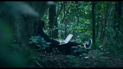 A person wearing all black sits at he base of a tree using a laptop in the middle of the forest.