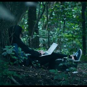 A person wearing all black sits at he base of a tree using a laptop in the middle of the forest. 