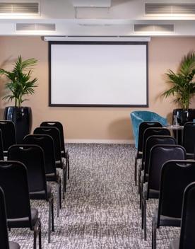 A conference room in the Rydges Hotel located at 75 Featherston Street, Pipitea in Wellington. The room has a grey carpet, beige walls, seven rows of black chairs facing the front of the room.