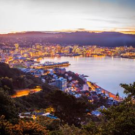 Looking down on Wellington city from Mount Victoria at sunset, the orange glow of the city lighting up is seen as well as the purple hills and blue water.