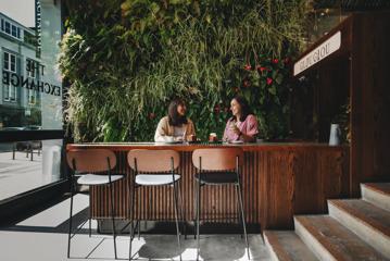 Two people sit at a bench in high chairs drinking coffee and eating decadent pastries. A green wall of plants is behind them and the words Glou Glou are above.