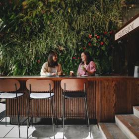 Two people sit at a bench in high chairs drinking coffee and eating decadent pastries. A green wall of plants is behind them and the words Glou Glou are above.