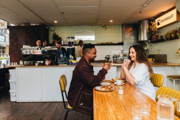 Two people sharing coffee and a meal inside Swimsuit on Dixon Street. The coffee counter is behind them with baristas working.