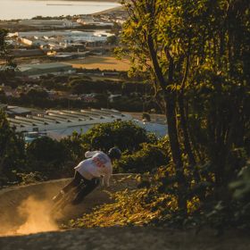A mountain biker riding along a dirt track amongst trees on Super Plus trail in Wainuiomata Park. In the background, there is a view of the Petone foreshore.