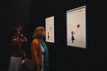 Three people look at screen prints displayed at The Art of Banksy exhibition.