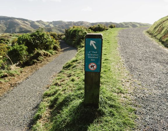 The top of the 4 degrees Track in Belmont Regional Park. A small signpost shows direction, with gravel paths cutting through farmland and hills in the background.