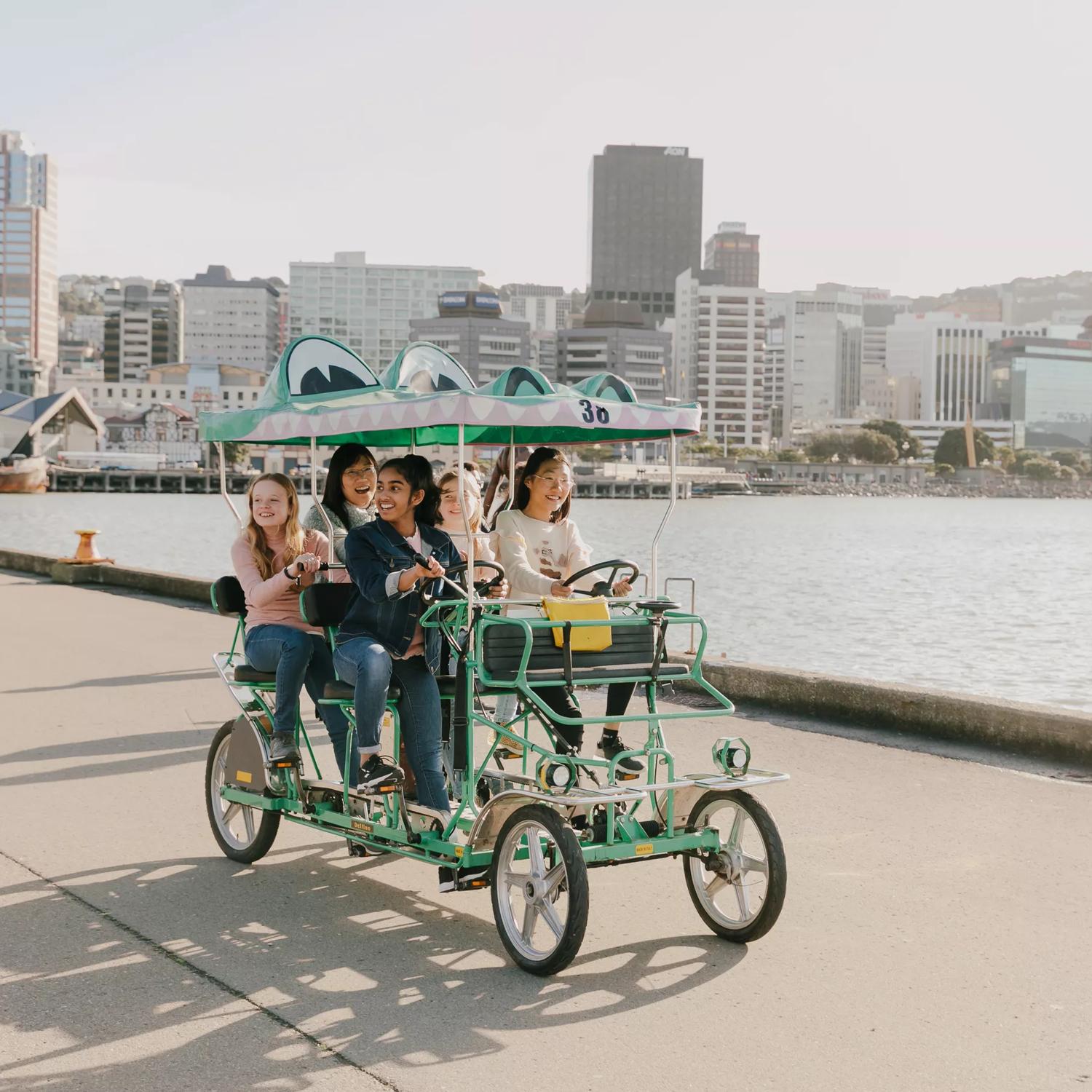 A group of teenagers riding a croc bike, a 5 seater, 4 wheeled green cart that looks like a crocodile, along the Wellington waterfront.
