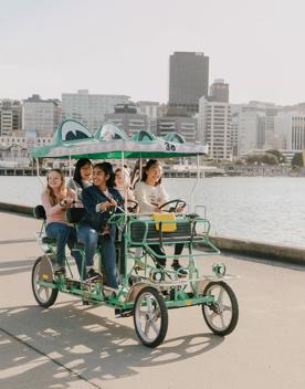 A group of teenagers riding a croc bike, a 5 seater, 4 wheeled green cart that looks like a crocodile, along the Wellington waterfront.