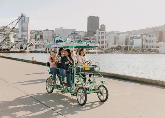 A group of teenagers riding a croc bike, a 5 seater, 4 wheeled green cart that looks like a crocodile, along the Wellington waterfront.