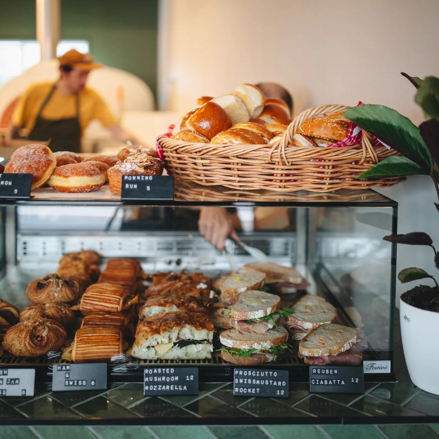 The front counter of a bakery with sandwiches and baked goods fo sale and two bakers working in the back.