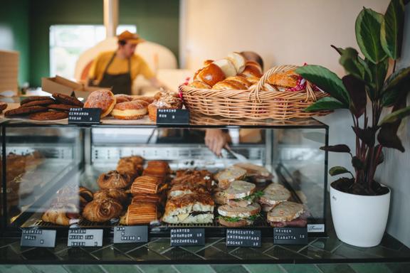 The front counter of a bakery with sandwiches and baked goods fo sale and two bakers working in the back.