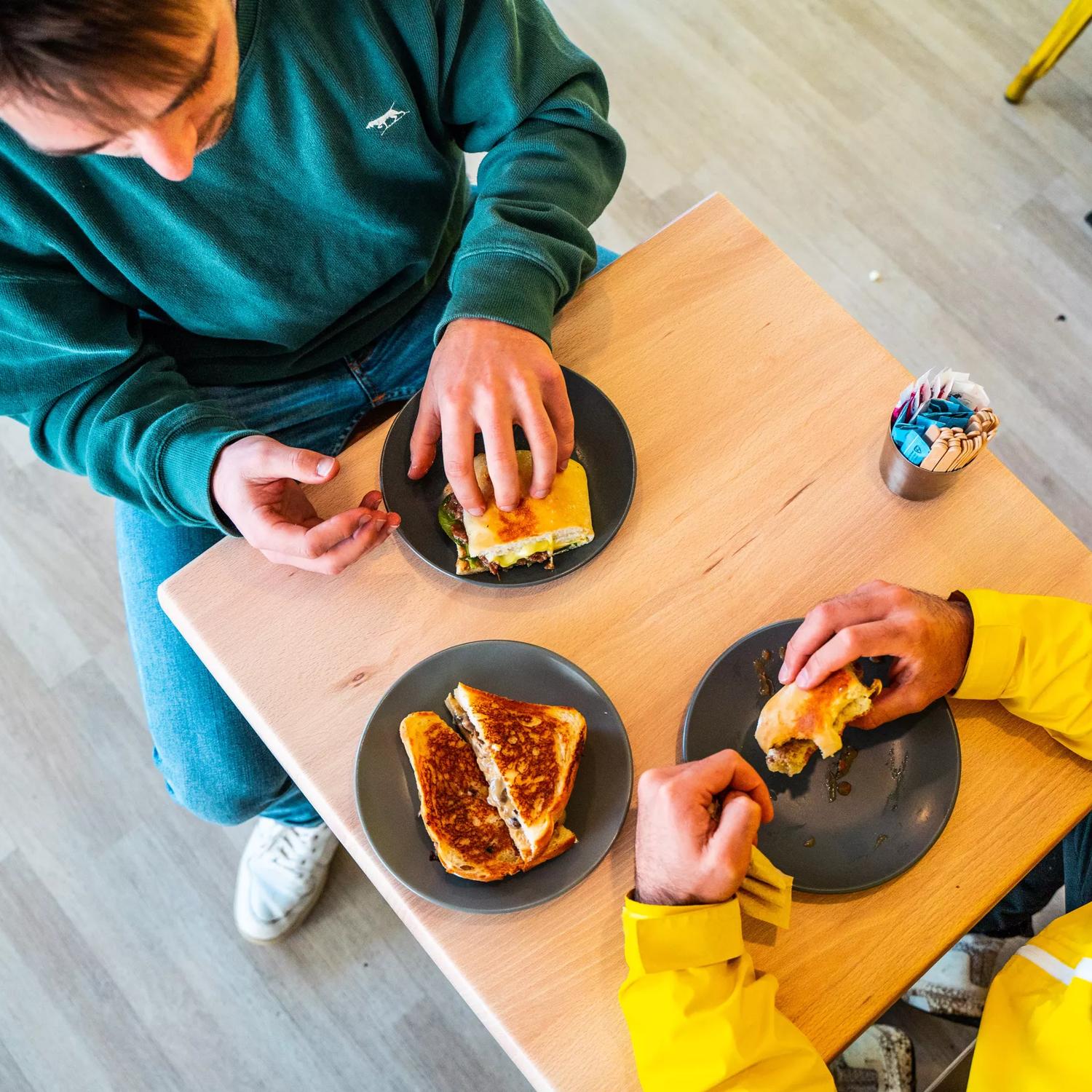 Shot from above, two people sit at a small square table eating sandwiches at Tricky's Sandwich Co., a sandwich shop in Upper Hutt, Hutt Valley.