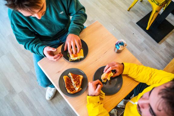 Shot from above, two people sit at a small square table eating sandwiches at Tricky's Sandwich Co., a sandwich shop in Upper Hutt, Hutt Valley.