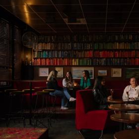 The interior The Library, a cocktail bar in Courtenay Place, Te Aro, Wellington. There are two groups of patrons drinking. The booths are red velvet and the walls are lined with books.