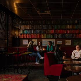 The interior The Library, a cocktail bar in Courtenay Place, Te Aro, Wellington. There are two groups of patrons drinking. The booths are red velvet and the walls are lined with books. 