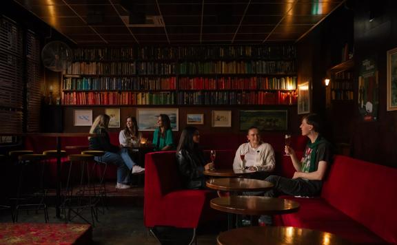 The interior The Library, a cocktail bar in Courtenay Place, Te Aro, Wellington. There are two groups of patrons drinking. The booths are red velvet and the walls are lined with books. 