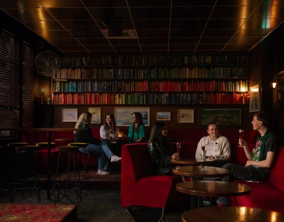 The interior The Library, a cocktail bar in Courtenay Place, Te Aro, Wellington. There are two groups of patrons drinking. The booths are red velvet and the walls are lined with books. 