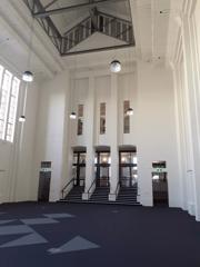 The Great Hall at Massey University in Mount Cook, Wellington. The large space is empty with a blue carpet and white walls. There are three doors on one side of the space with stairs leading up to them.