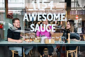 Three people sitting at a high table laughing behind a window at 1154 Pastaria. Text decal on the window reads “1154 Awesome Sauce”.