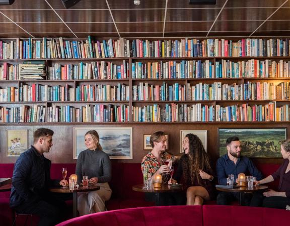 Six people sitting on tables in The Library, a cocktail bar and lounge on Courtenay Place.  The chairs are red velvet and the wall behind them is covered in books in bookshelves.