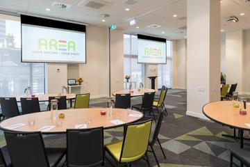 4 tables in conference room, each with 5 chairs, facing towards 2 projectors, both with AREAS's logo. notepads, pens, glasses, lollies and water pitchers are on the tables.