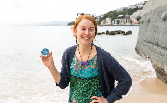 Natalie Jones, founder of Seasick Sunscreen Co, holds a tin of her company's product, smiling at Oriental Bay Beach in Wellington. 