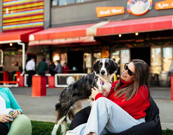 A person in a red sweater cuddles their dog sitting on a beanbag, on the grass at Glover Park, outside Rogue & Vagabond.